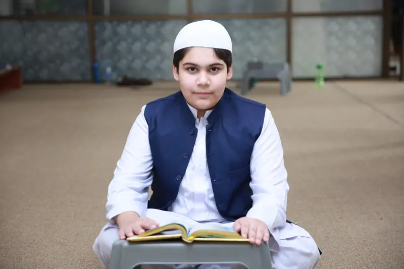 A student in academy uniform sitting cross-legged with a Quran open on a low desk