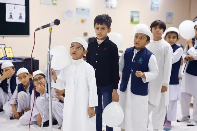 Students in white academy dress gathered indoors at an academy event, some holding white balloons