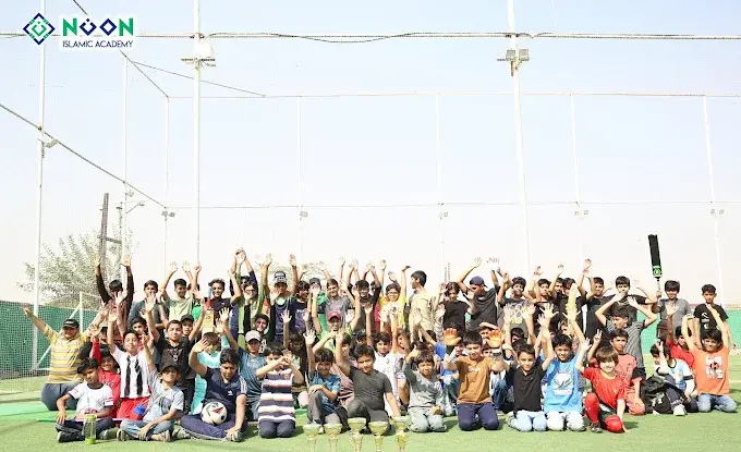 A large group of children cheering on an outdoor sports field, with trophies in front
